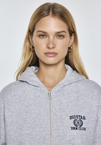 Vaaleanharmaa vetoketjullinen huppari, jossa on huppu, brodeerattu logo vasemmassa rinnassa, ribbaatut hihat ja pehmeä pinta. Varustettu hopeisella vetoketjulla.