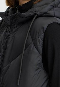 Schwarze Steppweste mit Kapuze, ausgestattet mit einem Rauten-Steppmuster, Reißverschluss und Zugbandsdetails. Glattes, leichtes Material.