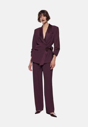 Blazer e pantaloni bordeaux in un tessuto morbido e strutturato. Il blazer presenta un profondo scollo a V, colletto a scialle e vita regolabile con una cintura.