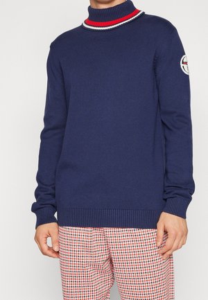 Mann trägt einen marineblauen Pullover mit rot-weiß gestreiftem Kragen und besticktem Logo am Ärmel, kombiniert mit rot-weiß gemusterten Hosen.