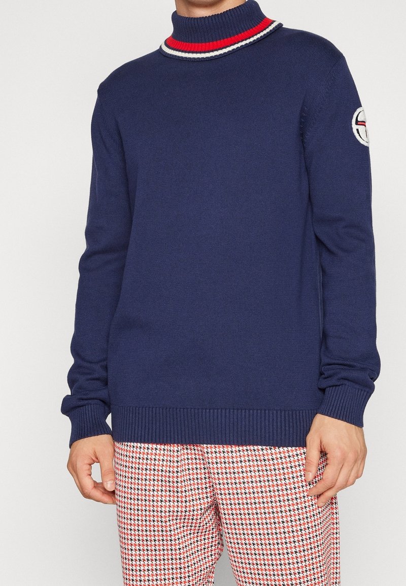 Homme portant un pull bleu marine avec un col rayé rouge et blanc et un logo brodé sur la manche, associé à un pantalon rouge et blanc à motifs.