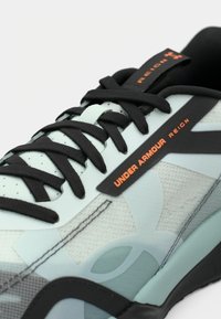 Αθλητικό παπούτσι με ανοιχτό πράσινο επάνω μέρος, μαύρα κορδόνια και λογότυπο Under Armour σε πορτοκαλί. Διαθέτει υφασμάτινα πάνελ και κομψό σχέδιο.