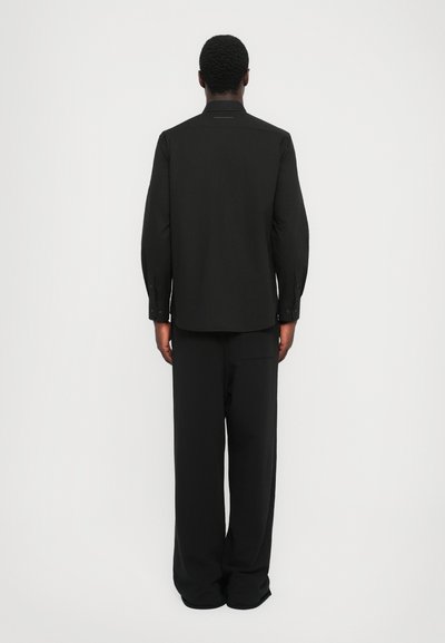 Chemise noire à manches longues avec une coupe droite, des poignets boutonnés et un col discret. Associée à un pantalon ample assorti.