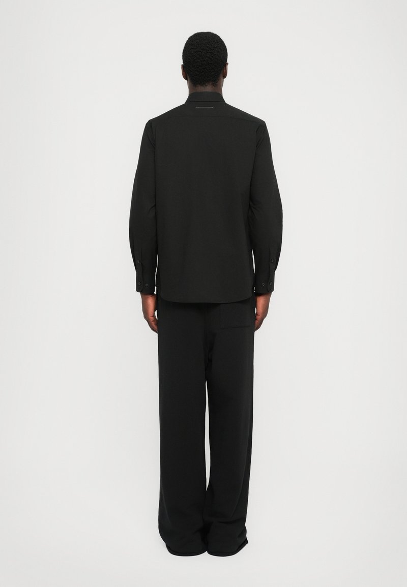 Sort skjorte med lange ærmer, lige snit, knaplukning ved ærmerne og en diskret krave. Sammen med matchende løse bukser.