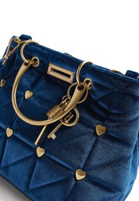 Sac à main matelassé en velours bleu avec ferrures dorées, présentant des accents en forme de cœur et un détail porte-clés. Forme structurée avec double poignée.