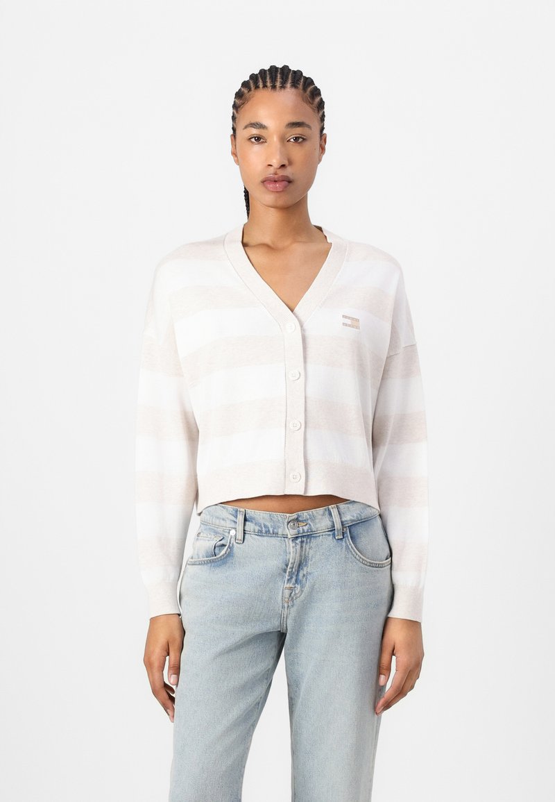 Cárdigan corto y ligero en beige con rayas blancas, con escote en V, cierre de botones y puños acanalados. Lleva puesto sobre jeans de un azul claro.