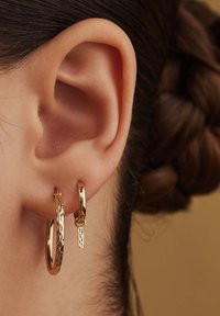 Goldene Creolen werden an einem Ohr präsentiert; eine große, glatte Creole und eine kleinere, strukturierte mit einem hängenden Charme für zusätzliche Details.