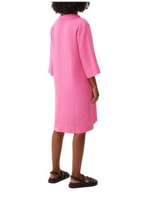 Rosa Leinenkleid mit Dreiviertelärmeln, Kragen, lockerer Passform und Oberschenkel-langer Länge, kombiniert mit schwarzen Sandalen mit Riemchen.