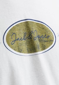 Tricou alb cu un grafic oval mare de culoare verde măslin și bleumarin, cu "Jack & Jones" într-un font cursiv. Textură moale din bumbac.