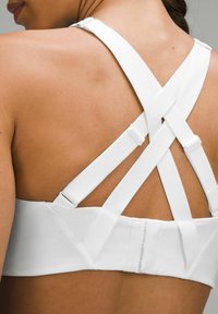 Sujetador deportivo blanco con tirantes anchos cruzados en la espalda, mostrando una textura suave y cierre de gancho y ojo. Diseño minimalista sin estampados.