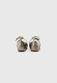 Sandales dorées métalliques avec bride de cheville, semelle plate, orteil arrondi, texture lisse et design minimaliste. Vue arrière mettant en avant le détail de la boucle.