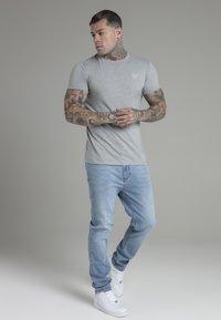 Grijze kortekleurige shirt met een logo op de borst, gecombineerd met lichtblauwe jeans en witte sneakers. Huidkenmerken tonen zichtbare tatoeages.