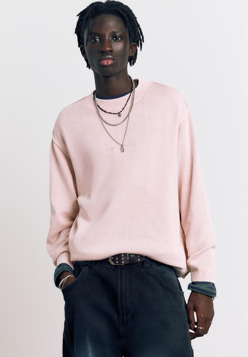 Jeune personne à la peau foncée et aux dreadlocks portant un pull rose clair, des colliers superposés et un pantalon sombre, les mains partiellement dans les poches.
