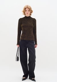 Top nero a maniche lunghe con un motivo testurizzato a pois dorati, abbinato a pantaloni in denim neri a gamba larga. Il modello tiene una piccola borsa nera.