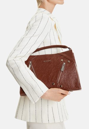 Mujer con blazer blanco de rayas finas sosteniendo un bolso de cuero marrón con detalles de cremalleras plateadas y asa superior.