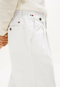 Pantaloni bianchi in cotone con pieghe, dotati di chiusura con bottone e tasche laterali. Piccolo logo del marchio in rosso, bianco e blu alla vita.