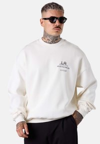 White sweatshirt with ribbed cuffs and neckline, featuring a black printed logo on the chest. Model wearing black sunglasses.