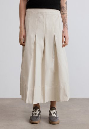 Plissierter, knöchellanger Rock aus hellbeigem Stoff. Verfügt über einen glatten Bund und einen asymmetrischen Saum. Kombiniert mit grauen Sneakers.