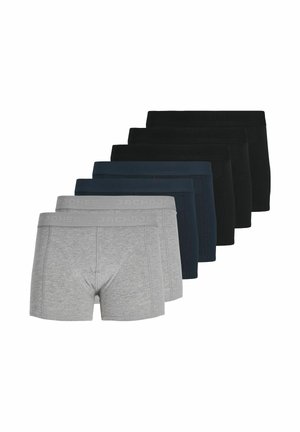 Młody mężczyzna z kręconymi brązowymi włosami, ubrany w dopasowane szare bokserki, stoi na jasnym, jednolitym tle.