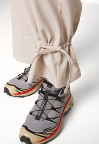 Μπεζ παντελόνια με κορδόνι στη μέση και συγκεντρωμένα μανίκια, σε συνδυασμό με γκρι αθλητικά παπούτσια Salomon που διαθέτουν μαύρες και κόκκινες λεπτομέρειες και υφή στην επιφάνεια.