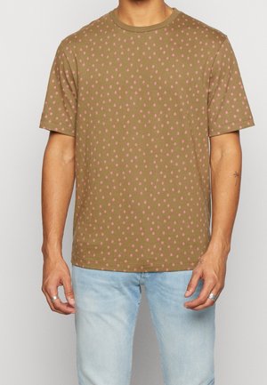 Hombre que lleva una camiseta de manga corta verde oliva con un pequeño patrón de rombos rosas y vaqueros azul claro, de pie con los brazos a los lados.