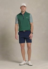Chaleco verde con cremallera sobre una polo de rayas, pantalones cortos navy y zapatos de golf blancos. Accesorios incluyen una gorra blanca y un reloj. Diseño bien ajustado.