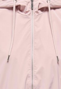 Gaiši rozā jaka ar centrālu sudraba rāvējslēdzēju un baltām auklām ar metāla galiņiem pie kapuces atvēruma.