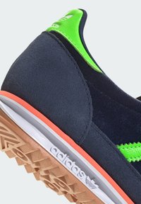 Sneaker din nubuc bleumarin cu o suprafață texturată, având accente verzi strălucitoare și o talpă intermediară portocalie. Logo debossat pe lateral.