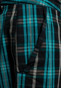 Tessuto a quadri nero e verde petrolio con motivo rettangolare, caratterizzato da strisce grigie. Dettaglio di tasca visibile con cuciture a contrasto.