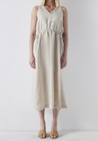 Robe maxi en lin beige avec encolure en V, taille ajustable par cordon, boutons sur le devant et poches latérales. Associée à des sandales à enfiler couleur beige sable.