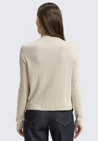 Kobieta o prostych brązowych włosach, ubrana w beżowy sweter z długim rękawem i ciemnoniebieskie dżinsy, stoi tyłem na neutralnym tle.