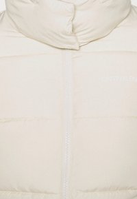 Gilet imbottito color crema con colletto alto, chiusura con zip e marchio discreto. Texture liscia con un motivo trapuntato per l'isolamento.