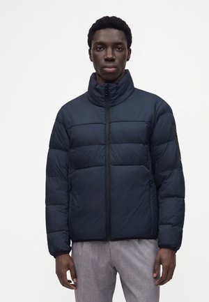 Homme portant une doudoune noire à col montant et fermeture éclair, debout devant un fond blanc uni, mains détendues le long du corps.