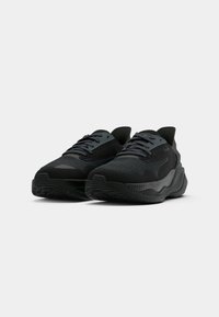 Paire de baskets de sport noires avec dessus en maille texturée et semelles épaisses et rembourrées sur un fond clair uni.