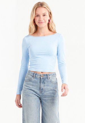 Ung kvinde med langt blond hår iført pink hoop-øreringe, en lyseblå cropped langærmet top og højttaljede lyseblå jeans.