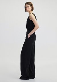 Černý overal s přiléhavým vrškem a širokými nohavicemi. Vyroben z hladkého materiálu, s tenkými ramínky a postranními kapsami, prezentovaný na světlejším pozadí.