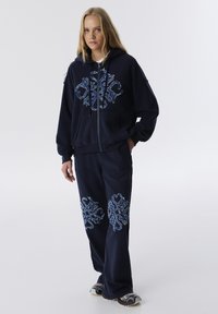 Marineblauwe rits-hoodie met een groot geborduurd patroon en een rafelige zoom, gecombineerd met bijpassende broeken met vergelijkbare borduursels op de onderbenen.