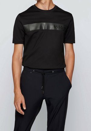 Mężczyzna w czarnej, krótkiej koszulce z błyszczącym poziomym paskiem i logo "BOSS", w połączeniu z czarnymi spodniami na sznurek.