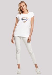 White cotton T-shirt with a colorful superhero logo in a diamond shape, paired with white skinny jeans and white sneakers.