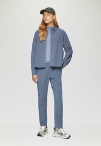 Lichtblauwe corduroyjas met knoopsluiting, gedragen over een lichtgrijze top, in combinatie met bijpassende broeken en zilveren sportschoenen.