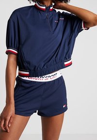 Marineblå cropped sportstop med halv lynlås, korte puffede ærmer og rød/hvide stribede detaljer. Samlet med matchende marineblå shorts.
