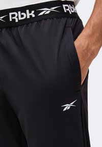 Gros plan d'une personne portant un short noir Reebok avec un logo blanc, une taille élastique et une main dans la poche latérale.