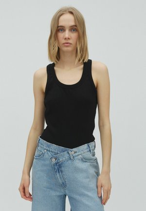 Jeune femme aux cheveux blonds porte un débardeur noir et un jean bleu clair avec la taille déboutonnée devant un fond gris uni.