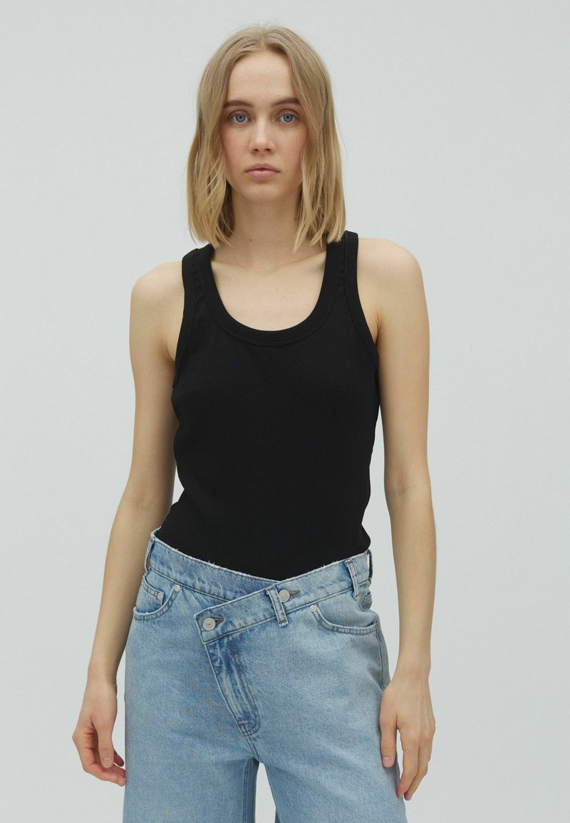 Jeune femme aux cheveux blonds porte un débardeur noir et un jean bleu clair avec la taille déboutonnée devant un fond gris uni.