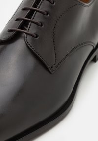 Chaussure habillée en cuir noir avec une texture lisse, design à lacets, accents cousus et une pointe arrondie. Semelle plate et style minimaliste.