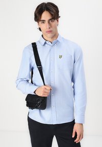 Chemise bleu clair à col boutonné avec une poche poitrine et un logo de papillon jaune. Le modèle porte un petit sac noir avec un accent en chaîne.