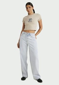Jovem mulher com cabelo trançado usa uma t-shirt curta bege da RVCA, jeans de pernas largas azul claro e sapatos pretos, de pé com a mão no bolso.