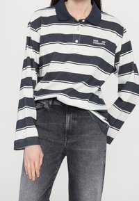 Polo a maniche lunghe bianco con righe orizzontali blu navy, con colletto blu navy e logo ricamato sul petto. Indossato con jeans grigi.