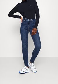 Höga, mörka denimjeans med en slim fit, kombinerat med en svart ribbad polotröja och vita sneakers med blå detaljer.