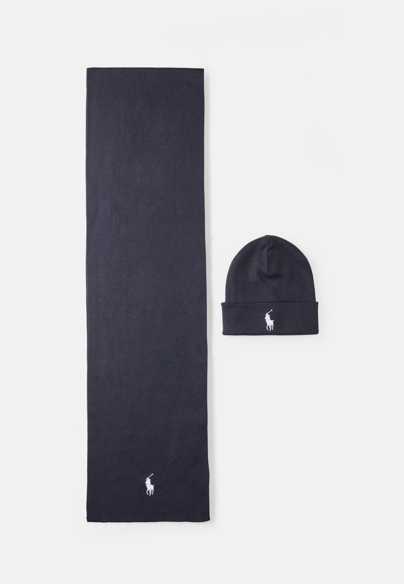 Mörkblå stickad halsduk och mössa uppsättning gjord av mjukt material, med en vit logotypdetalj på båda artiklarna. Halsduken är lång och rektangulär; mössan är åtsittande.